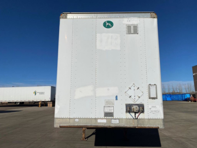 Rear view of a white Great Dane trailer with various stickers and fittings, including a green circular logo, vents, and electrical connections on the lower right; dockyard with other trailers in the background.