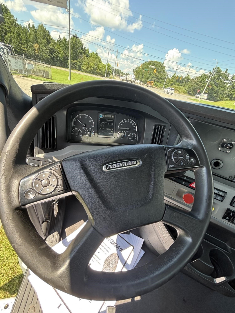 Steering wheel and dashboard of a Freightliner Cascadia day cab interior; visible instruments on the dash and a white device mounted near steering; view through windshield shows road.