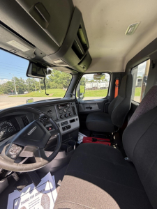 Truck cab interior showing steering wheel, dashboard, and front seats in a Freightliner Cascadia, with steering column controls and instrument cluster visible.