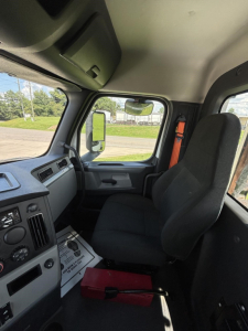 Truck cab interior showing driver's seat and dashboard of a Freightliner Cascadia; view through windshield and side mirror.