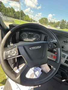 Steering wheel and dashboard of a Freightliner Cascadia day cab interior; visible instruments on the dash and a white device mounted near steering; view through windshield shows road.
