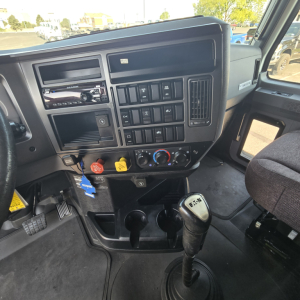 Truck cab interior showing dashboard with multiple switches, knobs, and a gear shifter. Center console includes a radio head unit, cup holders, and ignition area. Keys hanging from a blue tag.