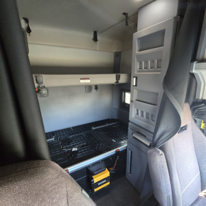 Truck cab interior showing sleeper area with overhead bunk and storage compartments, Mack ANTHEM dash area not visible, cargo area with grid floor and yellow toolbox.