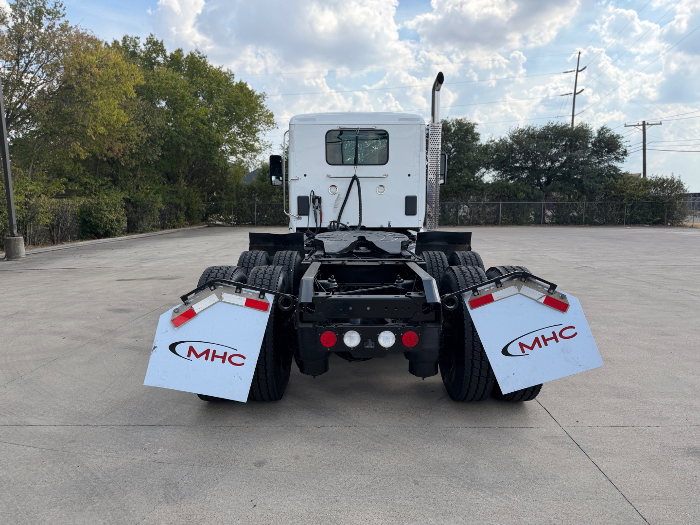 Kenworth T880 rear view showing dual rear wheels and mud flaps with MHC logos, white sleeper cab, and trailer hitch area.