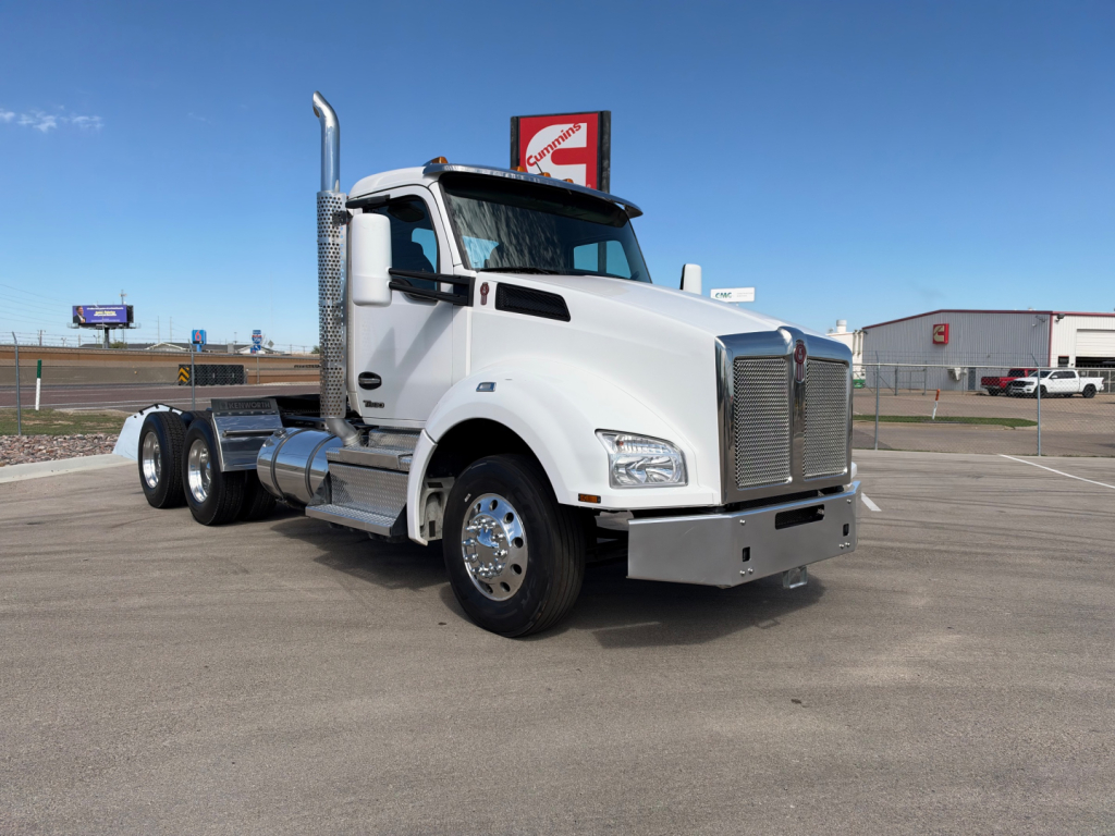 White Kenworth T880 truck with chrome exhaust stacks and grille, parked in lot. Visible text includes Kenworth emblem on grille and a sign in background.