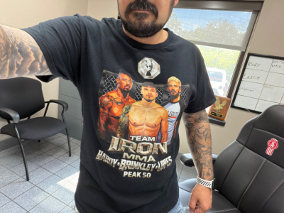 Image of a person taking a selfie inside a showroom with a 2022 Kenworth T880 listing note in background. Visible text on shirt: TEAM IRON MMA and various fighters. No vehicle details visible.