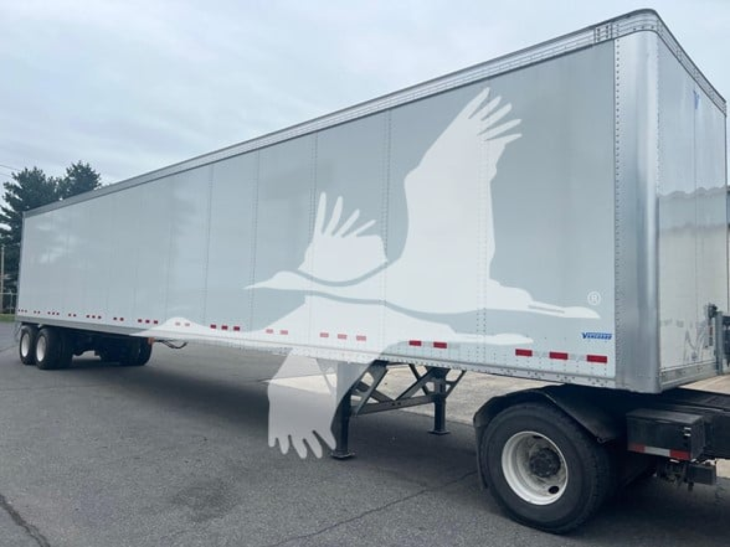 Trailer side view showing a Vanguard VXP Plate Van with a large white bird logo on the silver trailer body, dual rear wheels, and a front support stand.