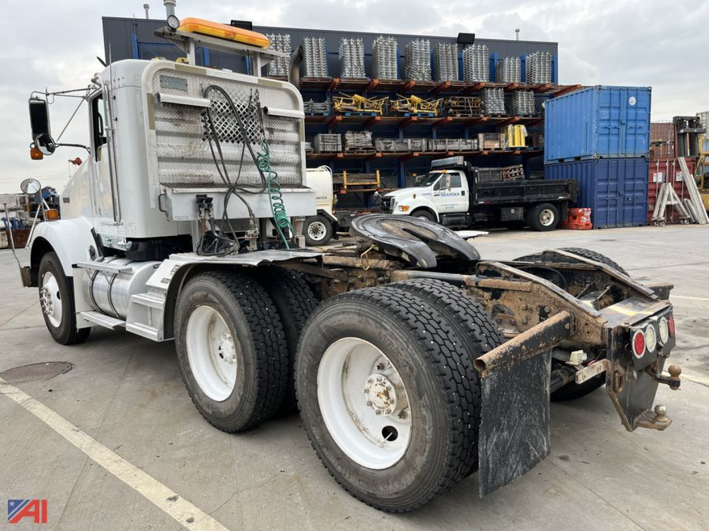 Kenworth T800 day cab tractor rear and dual rear axles with fifth wheel and mud flaps visible, industrial yard background with stacked pallets and shipping containers.