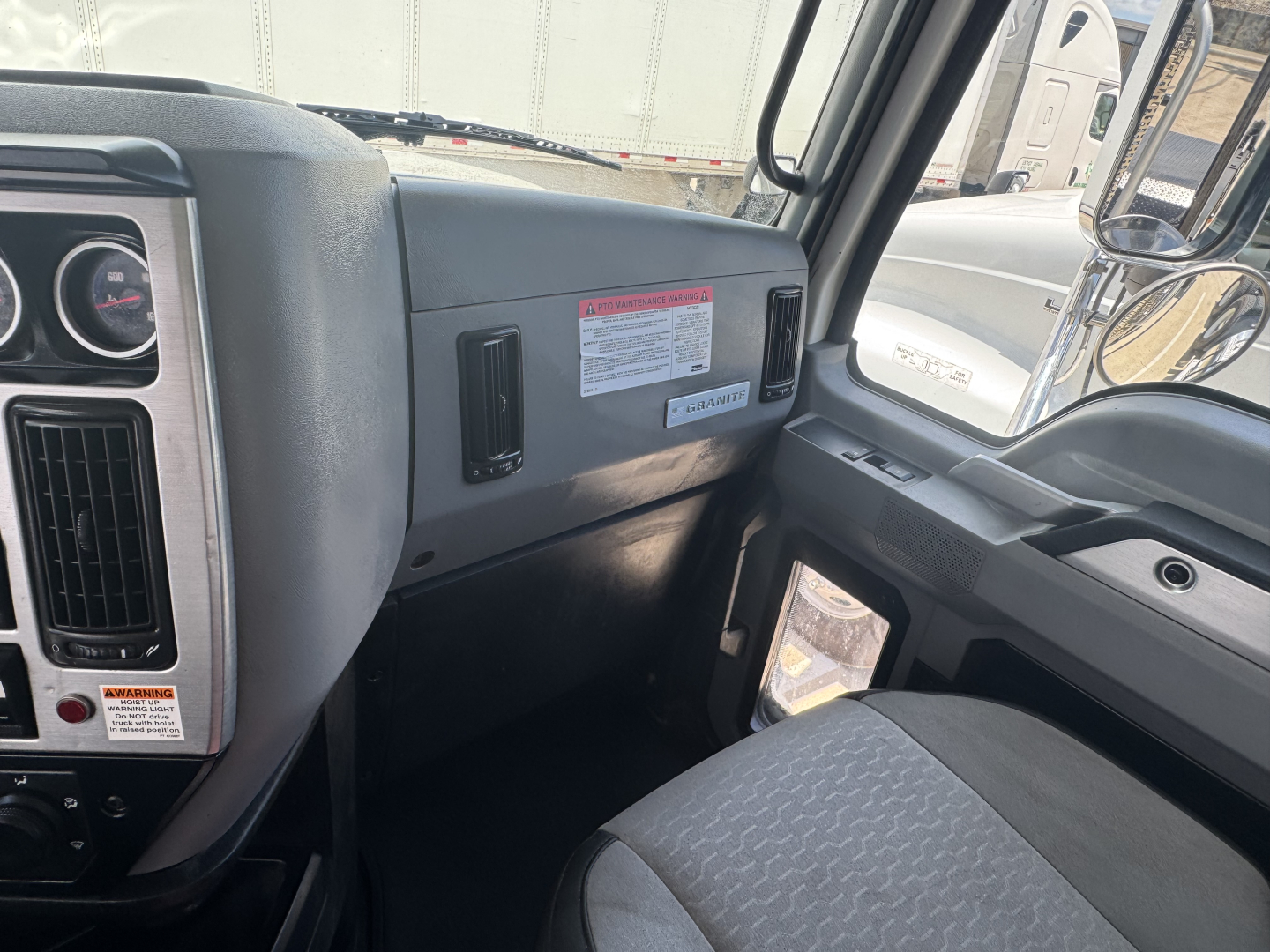 Cab interior of a 2023 Mack GR64B rolloff truck showing dashboard, steering wheel, driver door, and front passenger area with a warning label and vents.