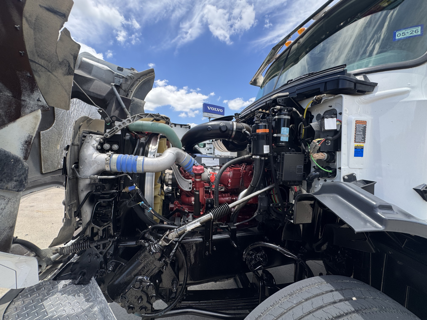 Truck engine bay of a 2023 Mack GR64B, showing engine, hoses, radiator, and various components in front of cab.