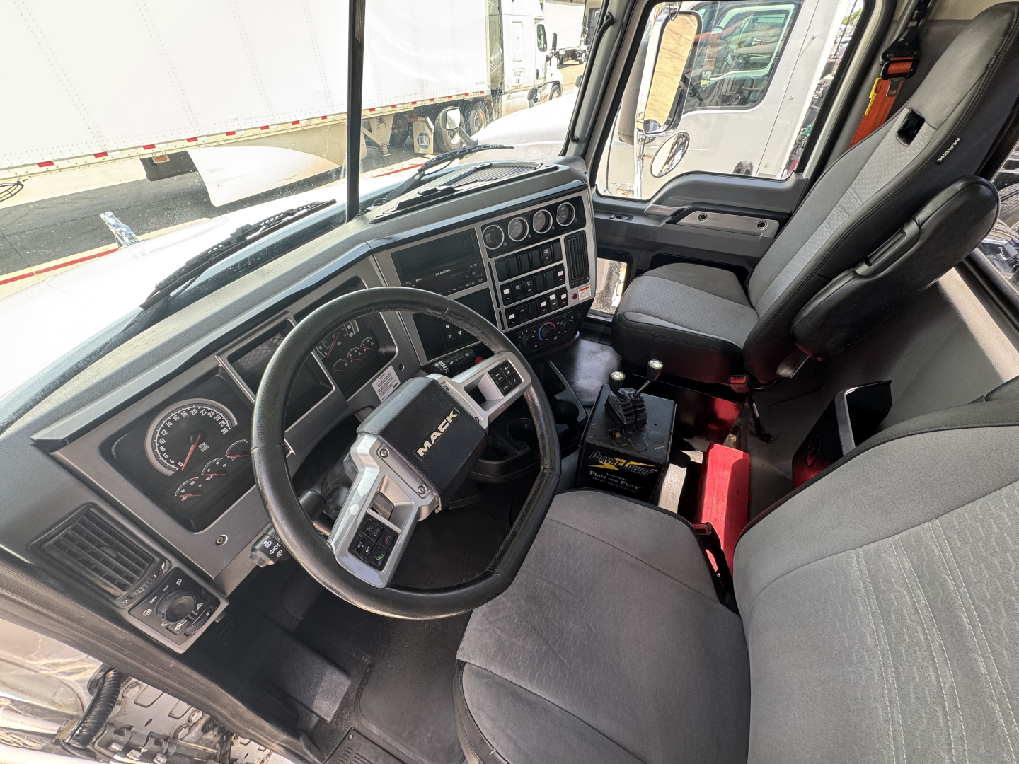 Cab interior of a Mack GR64B truck showing steering wheel, dashboard with gauges, gear shifter, driver and passenger seats, and center console.