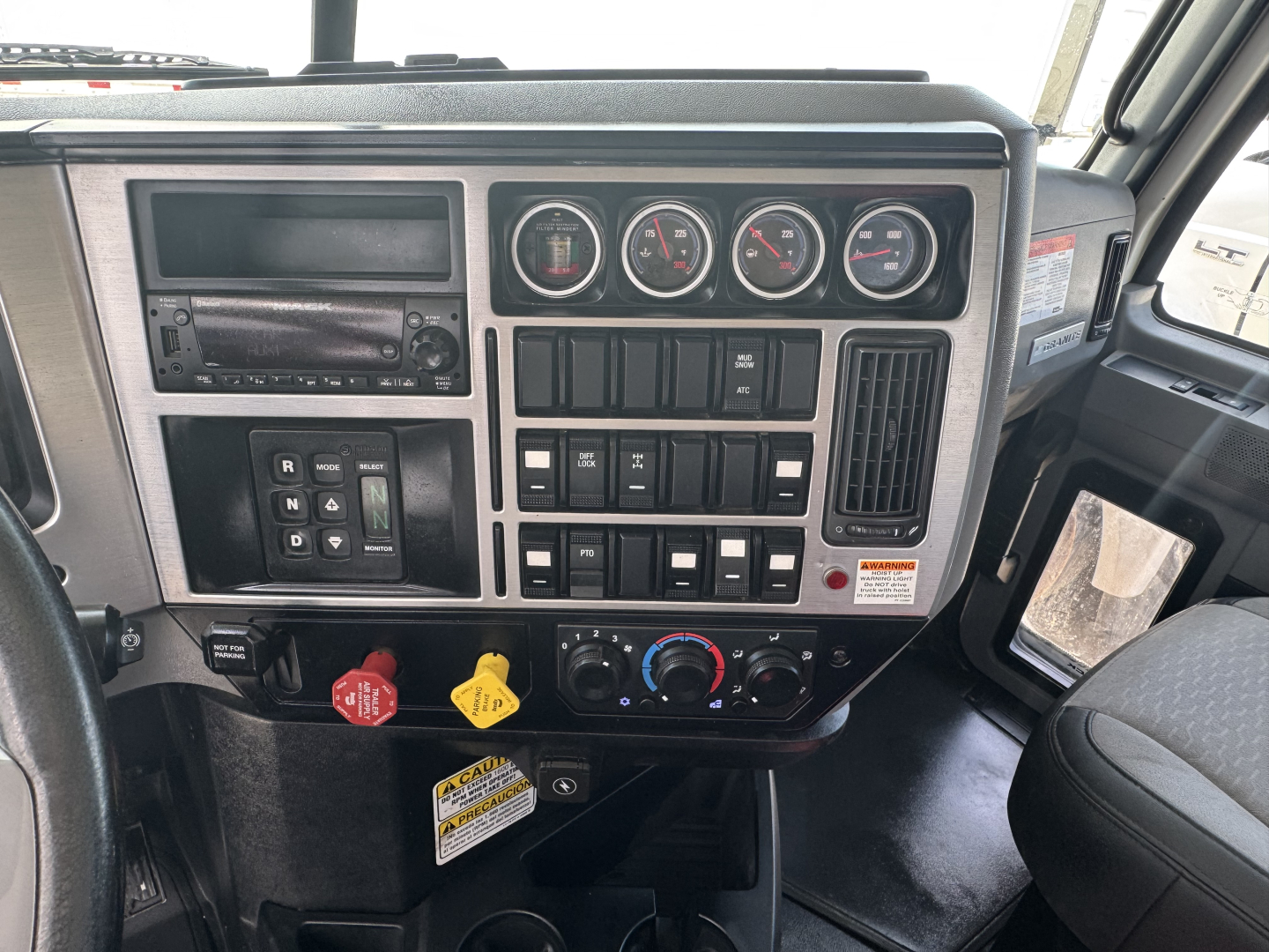 Dashboard panel of a Mack GR64B roll-off truck showing gauges, switches, climate controls, and radio in a metallic-framed cluster.