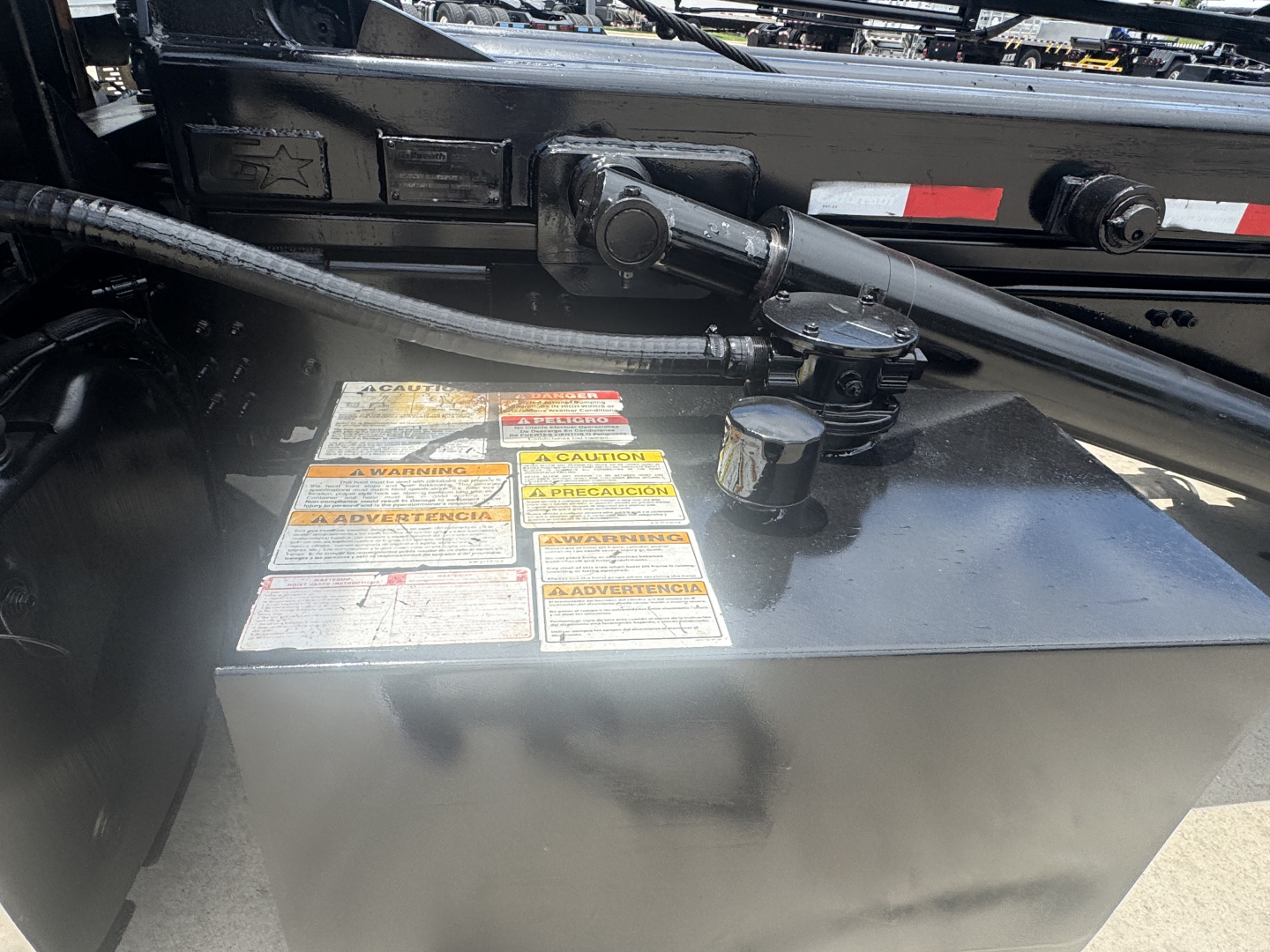 Trucks engine bay showing a metal rectangular fuel or battery box with warning labels on top and hydraulic hoses nearby.