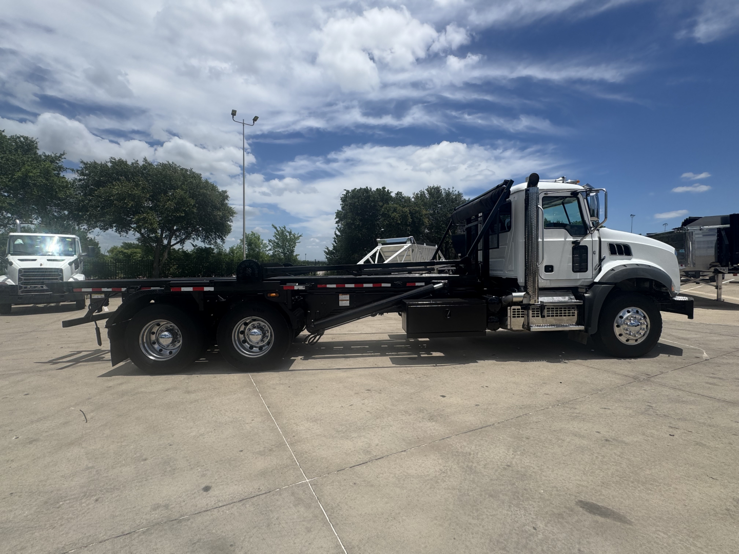 Flatbed/roll-off truck side view showing Mack GR64B cab and long equipment bed with dual rear axles and chrome rims.