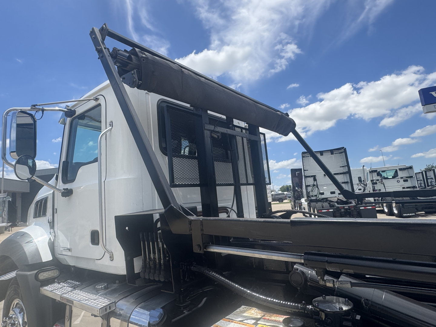 Mack GR64B roll-off truck cab in view showing hydraulic bed components and side steps.
