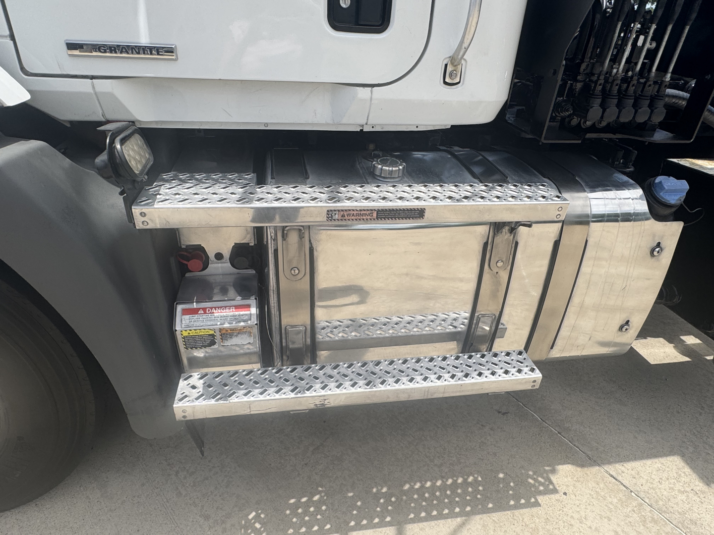Truck side step/footboard with aluminum diamond plate and fuel tank; Mack badge visible on cab; exposed mechanical components in a roll-off truck.