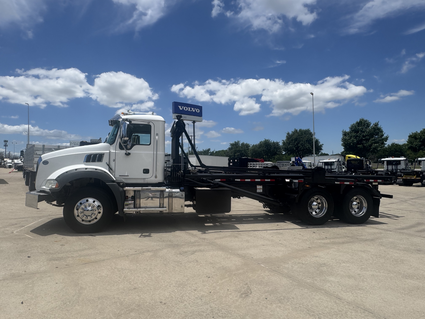 Truck side view of a white 2023 Mack GR64B roll-off with flatbed and rear axles visible, showing cab, long wheelbase, and chrome wheels.