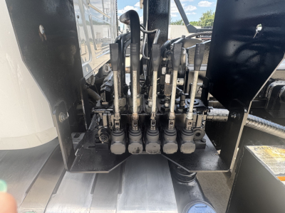 Six hydraulic levers and hoses in a Mack GR64B roll-off truck’s hydraulic control valve manifold, with metal rails and a warning label nearby.