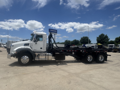 Truck side view of a white 2023 Mack GR64B roll-off with flatbed and rear axles visible, showing cab, long wheelbase, and chrome wheels.