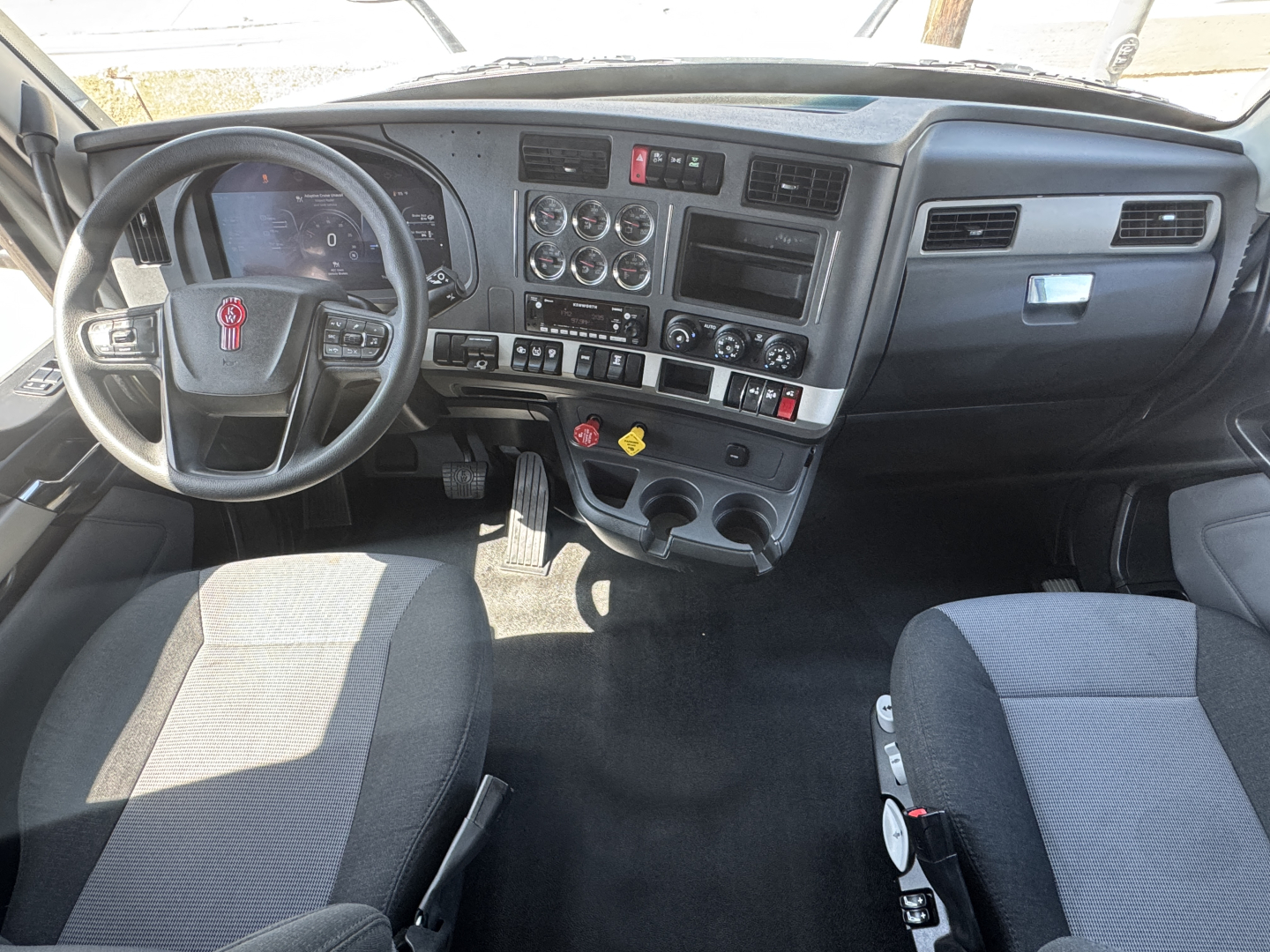 Kenworth T680 cab interior showing steering wheel with Kenworth emblem, dashboard with gauges, center console with knobs, cup holders, and gear shifter.