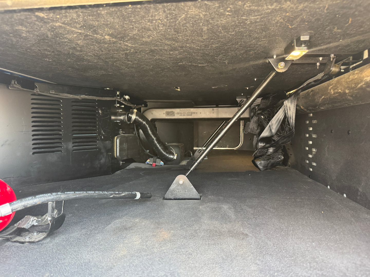 Kenworth T680 underbody storage area showing hydraulic lines and support strut with toolbox area. Text not legible.