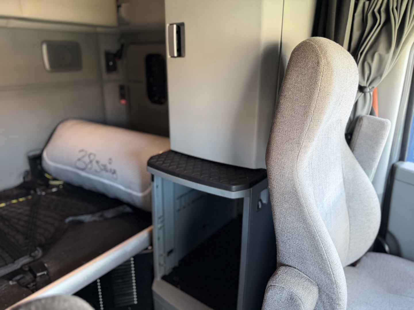 Truck cab interior showing driver's seat and sleeper area with storage cubby and dashboard area; silhouettes of curtains and window area visible.