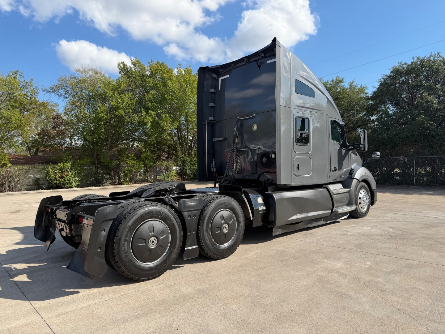 Truck tractor in a yard showing cab and drivetrain from rear-left side, 2022 Kenworth T680, dark gray color, 6x4 configuration, sleeper cab, no trailer attached.