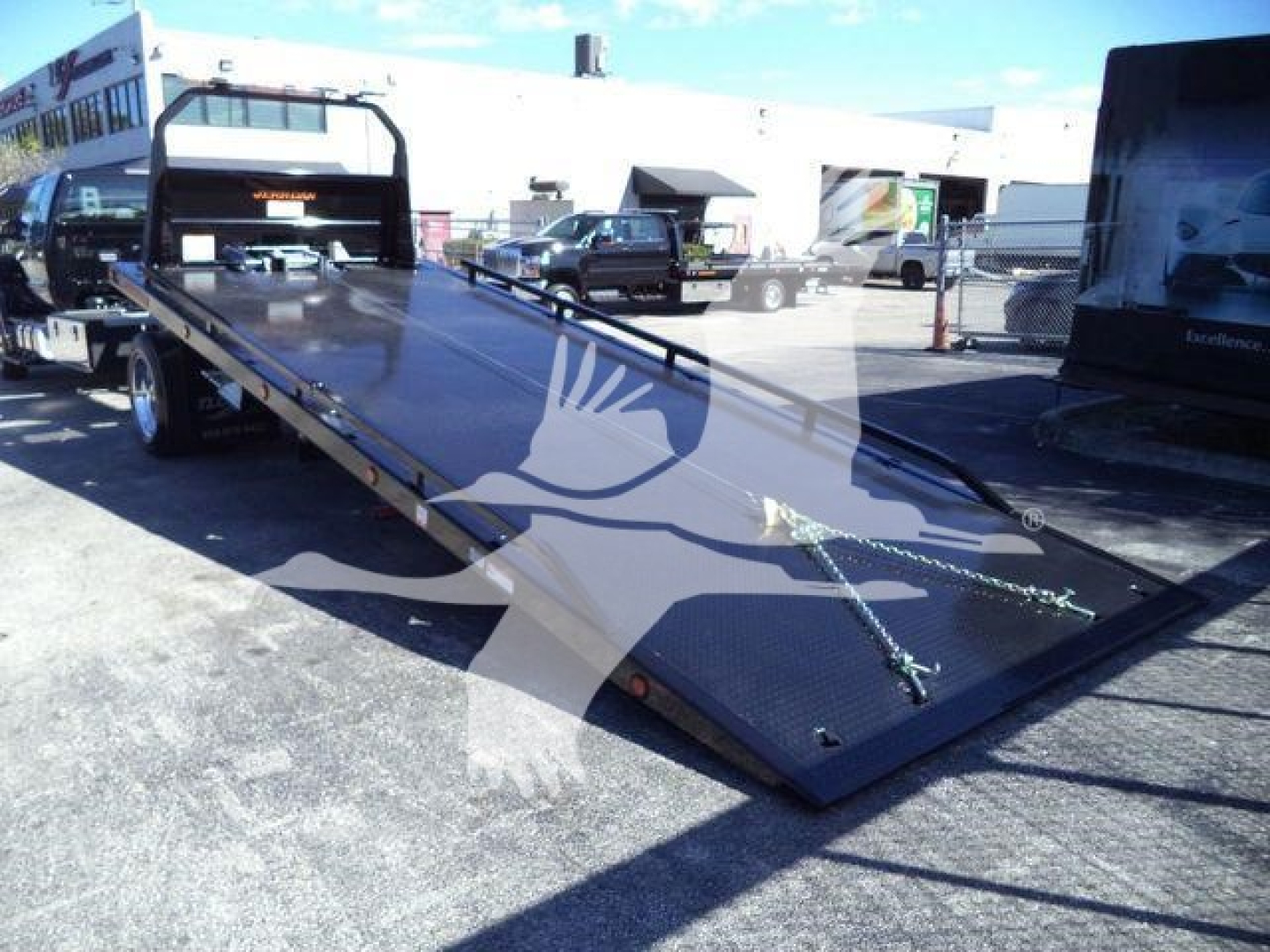Flatbed tow truck bed on a Ford F-750 rollback with ramp down, chains secured, urban lot in background.