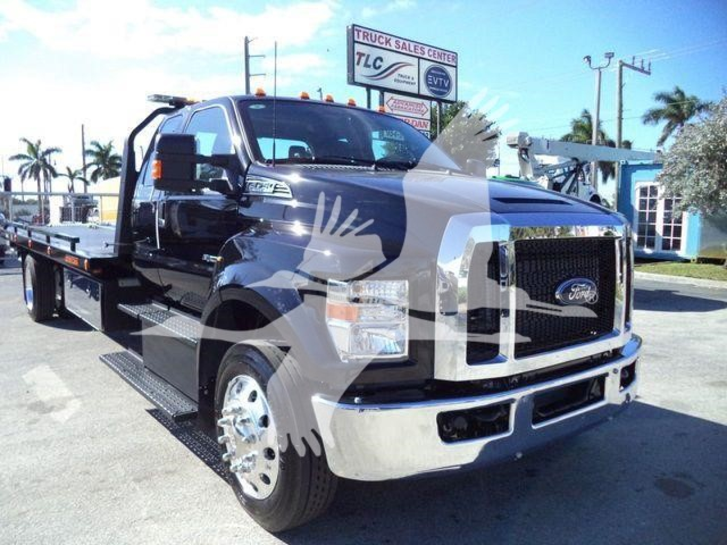 Ford F-750 tow truck with flatbed, black cab and chrome bumper, front grille visible, dealership in background.