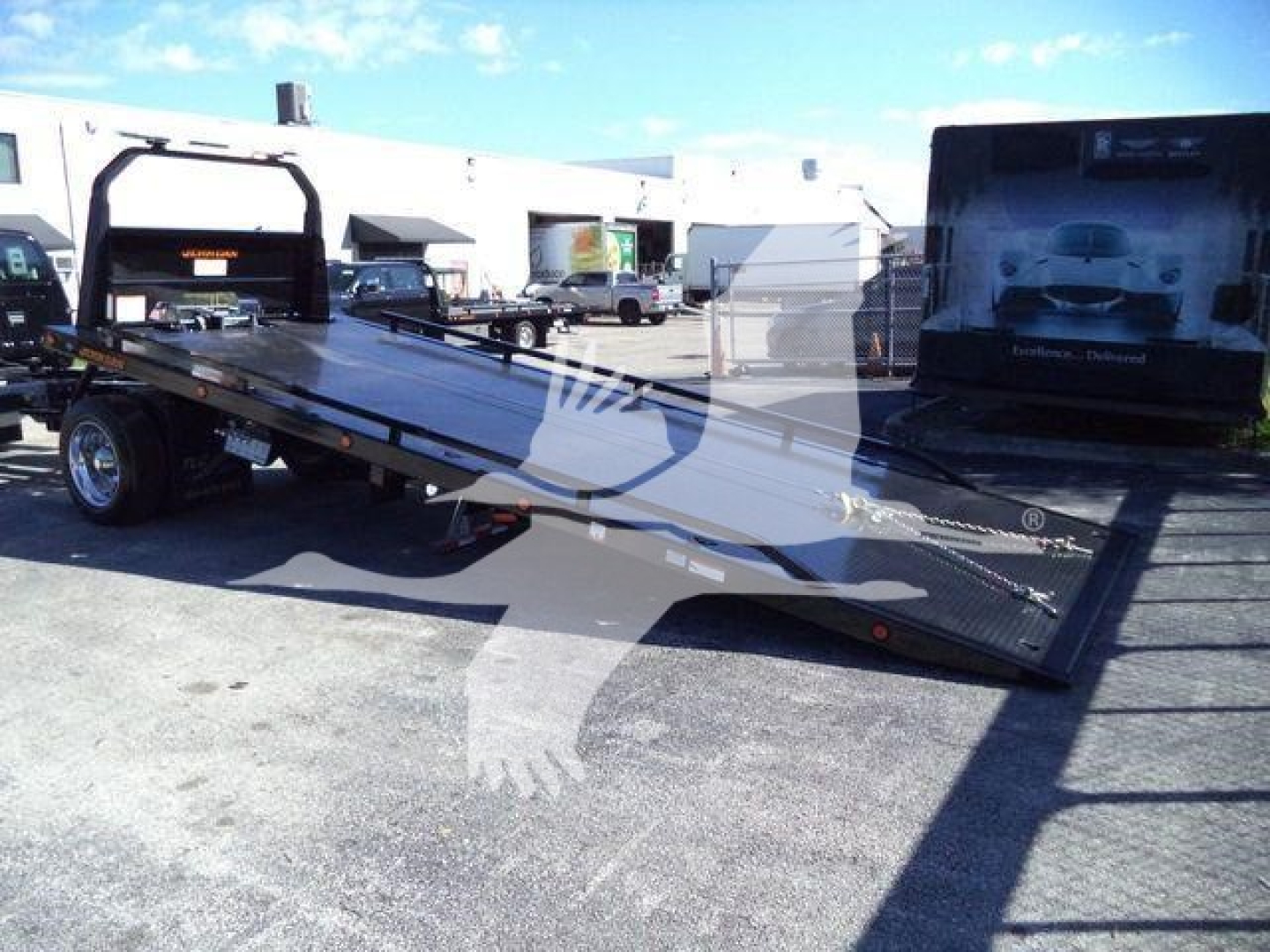 Ford F750 rollback tow truck flatbed with ramp extended, side rails, and wheel set visible; background shows dealership lot and other trucks.
