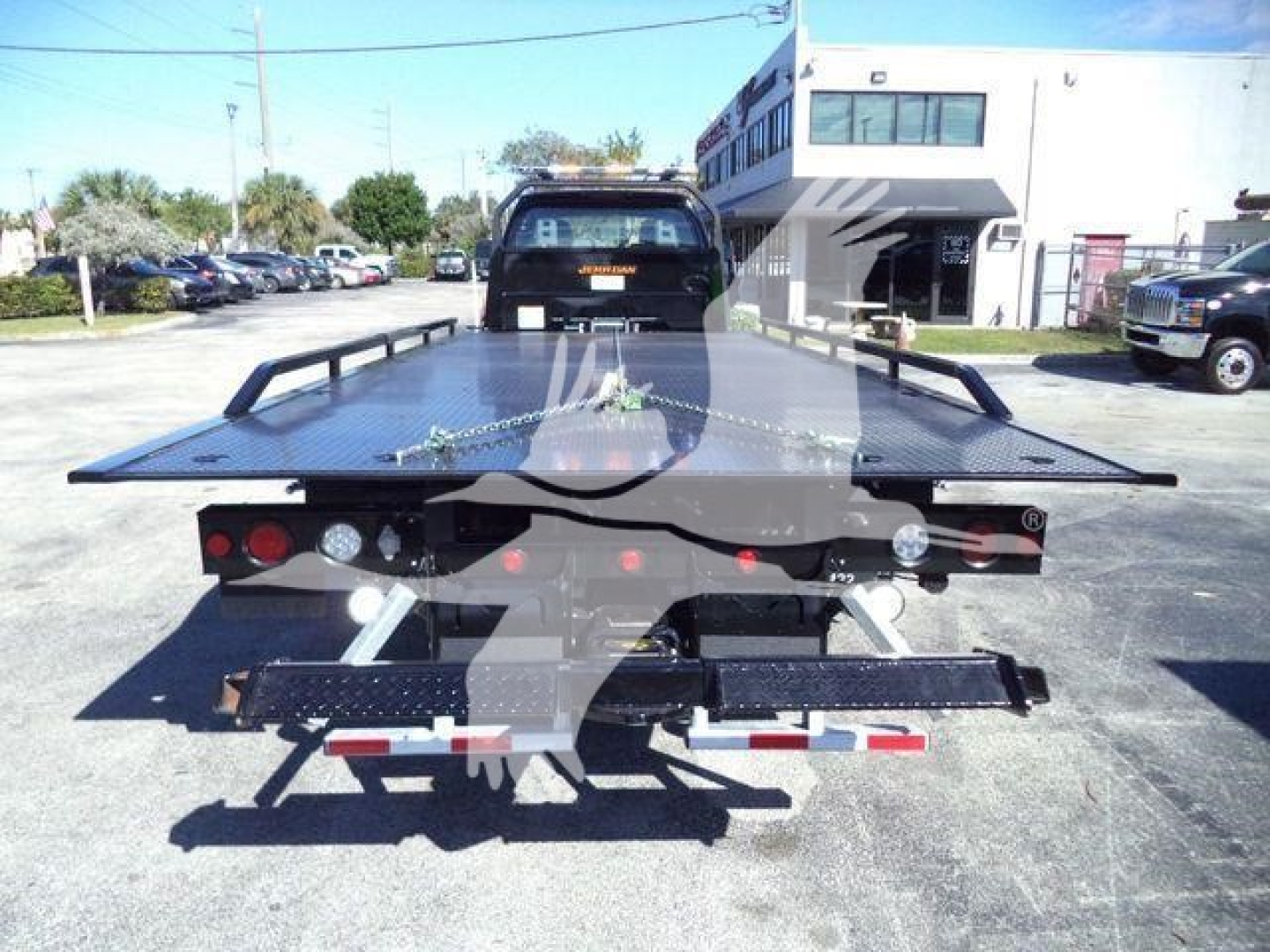 Flatbed rollback truck seen from rear, black platform with rails, tail lights, and towing equipment; text not clearly legible from distance.