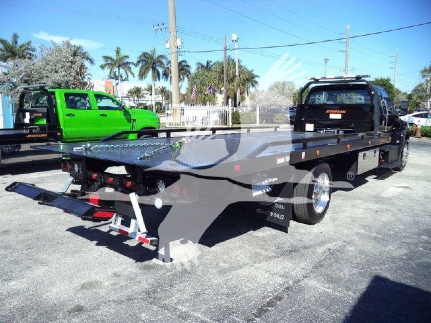 Flatbed tow truck trailer with black cab and green pickup in background, rear view showing loading ramps and hitch assembly.