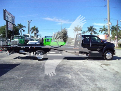 2024 Ford F750 tow truck flatbed side view showing cab and long bed with wheels, dealership lot in background