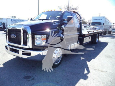 2024 Ford F750 Rollback truck chassis showing front grille with Ford badge and flatbed deck, in-service label visible on side window.