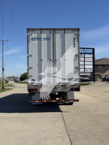 Rear of a white trailer with doors, showing model number 4044 and dealer logos, with a street and sign in background.