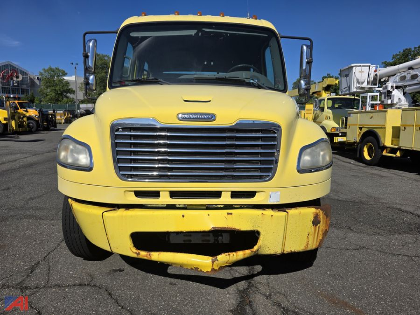 Freightliner M2 106 flatbed truck front view showing yellow cab and grille with bumper; service trucks in background.