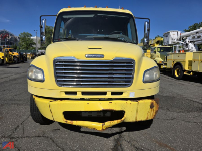 Freightliner M2 106 flatbed truck front view showing yellow cab and grille with bumper; service trucks in background.