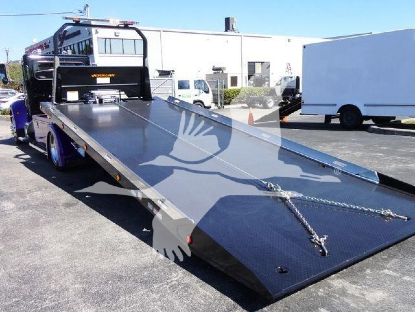Peterbilt 337 rollback truck with a long flatbed and raised gooseneck, black bed surface, towing chains, and cab in the background.