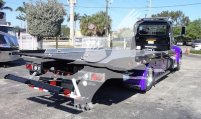 Truck bed of a 2022 Peterbilt 337 rollback w/ flatbed and rear lighting; visible label behind cab, purple accents on rear fenders; rear bumper and taillights visible.