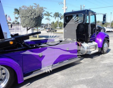 Peterbilt 337 rollback truck side view showing purple and black cab with chrome fuel tank and wheel wells, hydraulic boom mounting behind cab, roadside setting.