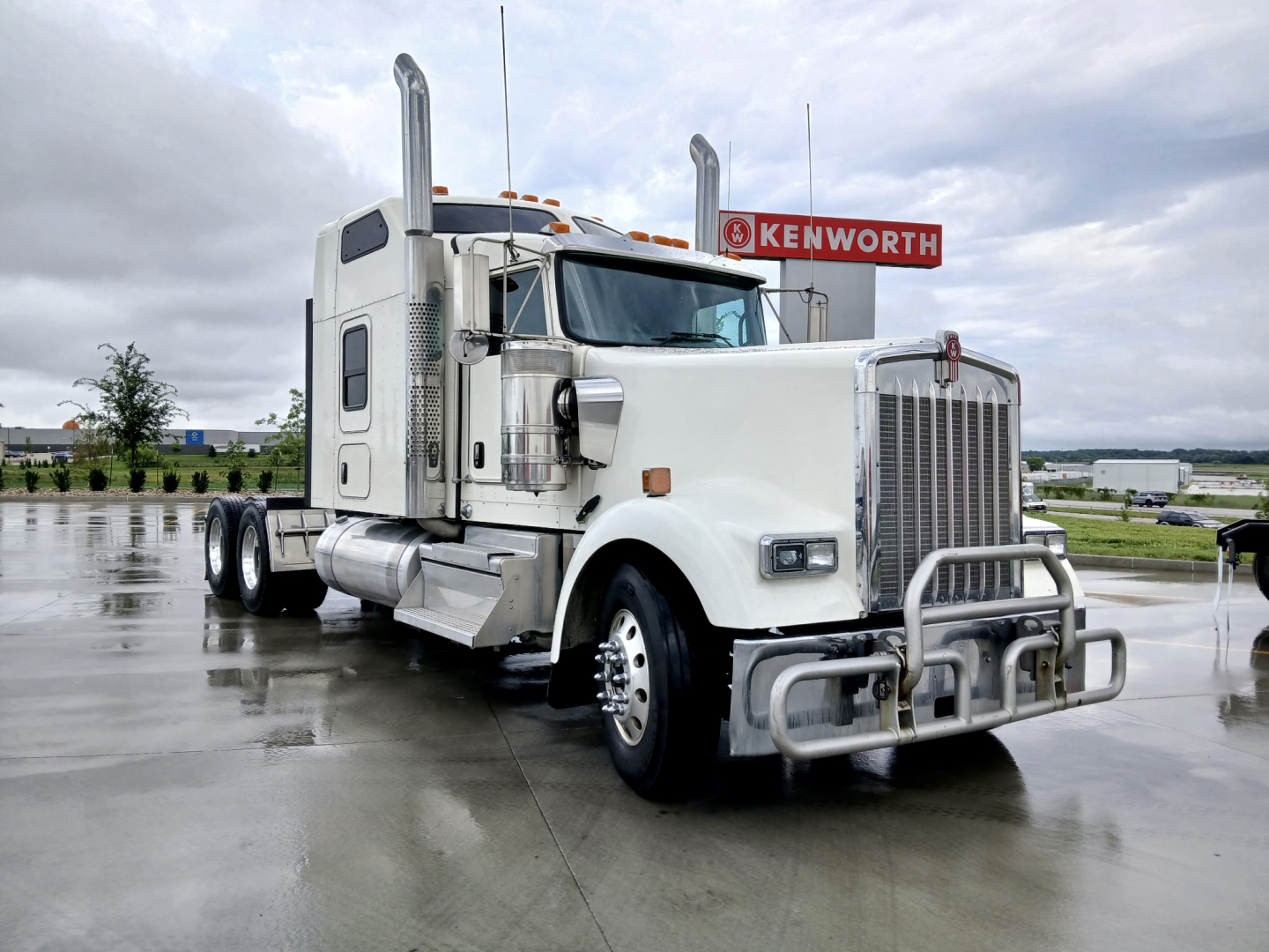 Kenworth W900L conventional sleeper truck in a parking lot, white cab with chrome grille and bumper, dual exhaust stacks, Kenworth sign in background.
