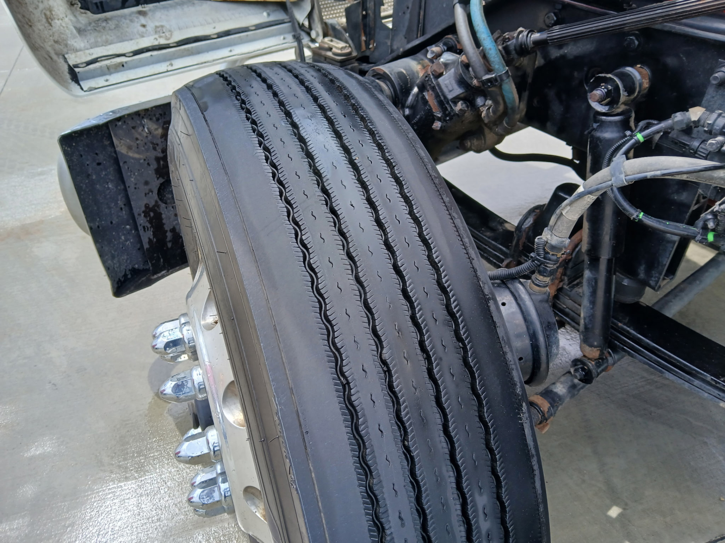 Kenworth W900L front axle area showing dual tandem wheel with treaded tire, hub, and suspension components.