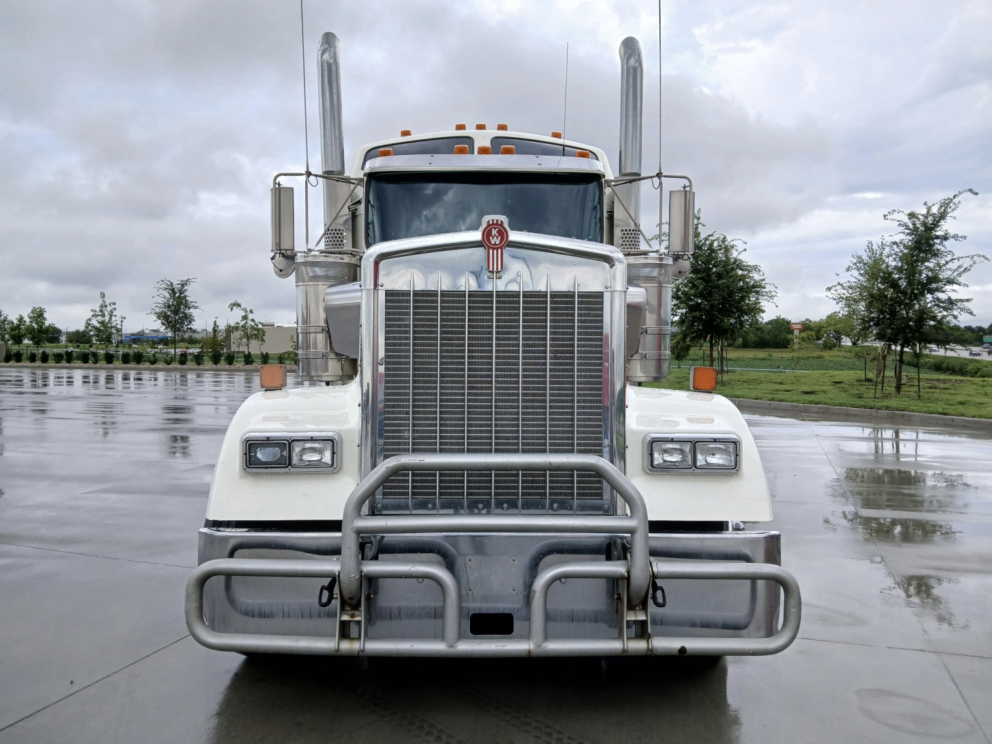 Kenworth W900L truck front view with tall exhaust stacks and chrome grille guard in a wet lot.