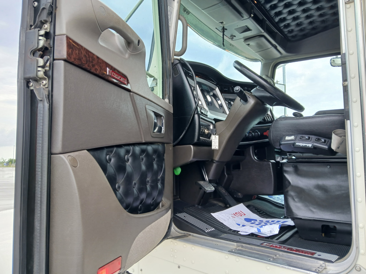 Kenworth W900L truck interior door panel and dashboard area visible; beige and brown interior; step plate; driver seat area; partial floor mats; dashboard with steering wheel