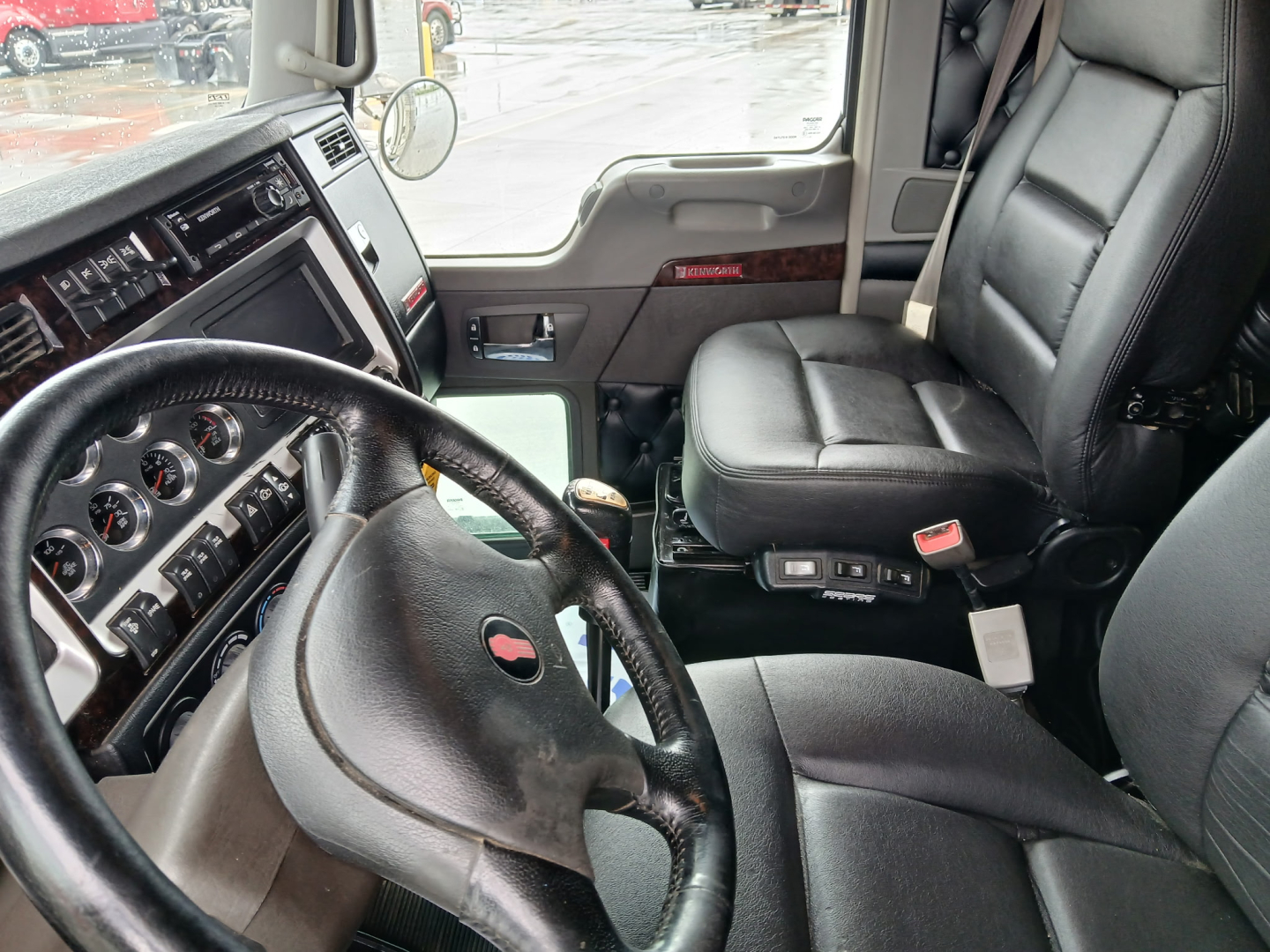Kenworth W900L interior showing steering wheel, dashboard with gauges, driver seat, and center console; black leather seats; door panel with wood trim.