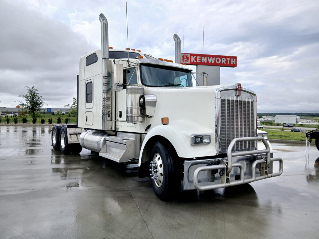 Kenworth W900L conventional sleeper truck in a parking lot, white cab with chrome grille and bumper, dual exhaust stacks, Kenworth sign in background.