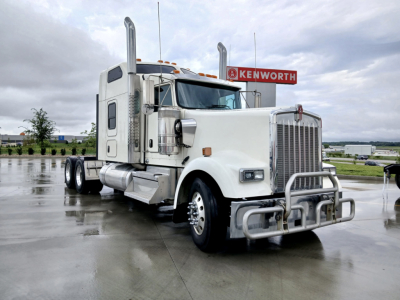 Kenworth W900L conventional sleeper truck in a parking lot, white cab with chrome grille and bumper, dual exhaust stacks, Kenworth sign in background.