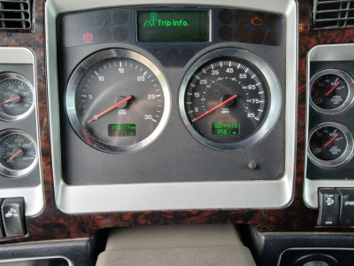 Dashboard of a 2023 Kenworth W900L showing two large gauges and a digital trip info screen in the center, with woodgrain trim surrounding the cluster.