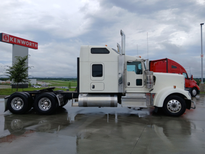 Kenworth W900L highway tractor side view with sleeper cab, fuel tanks, and chrome exhaust stacks at a dealership lot.