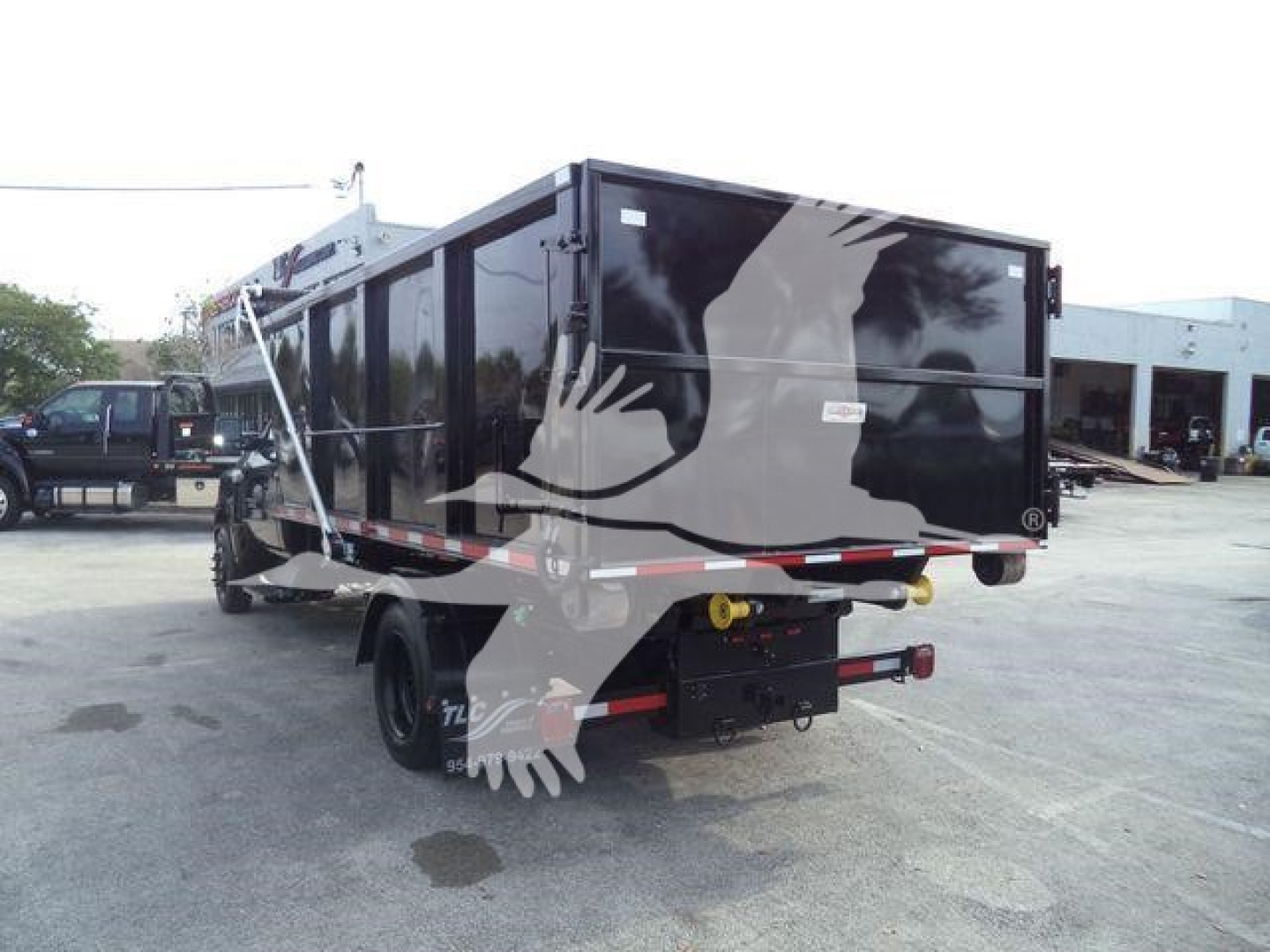 Truck flatbed trailer in a yard showing a black roll-off container mounted on a Silverado chassis with rear bumper and hydraulic hookups visible.